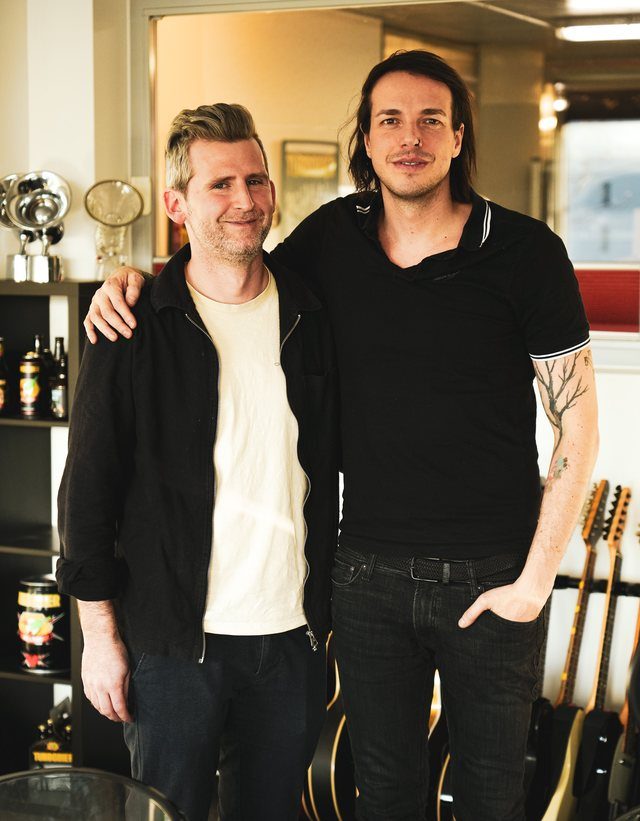 Michael Plank mit einem Gast mit langen Haaren, dahinter Gitarren